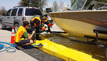 Photo of two provincial staff members in Selkirk, Manitoba, decontaminating the underside of a boat by spraying it with water at 60 degrees Celsius