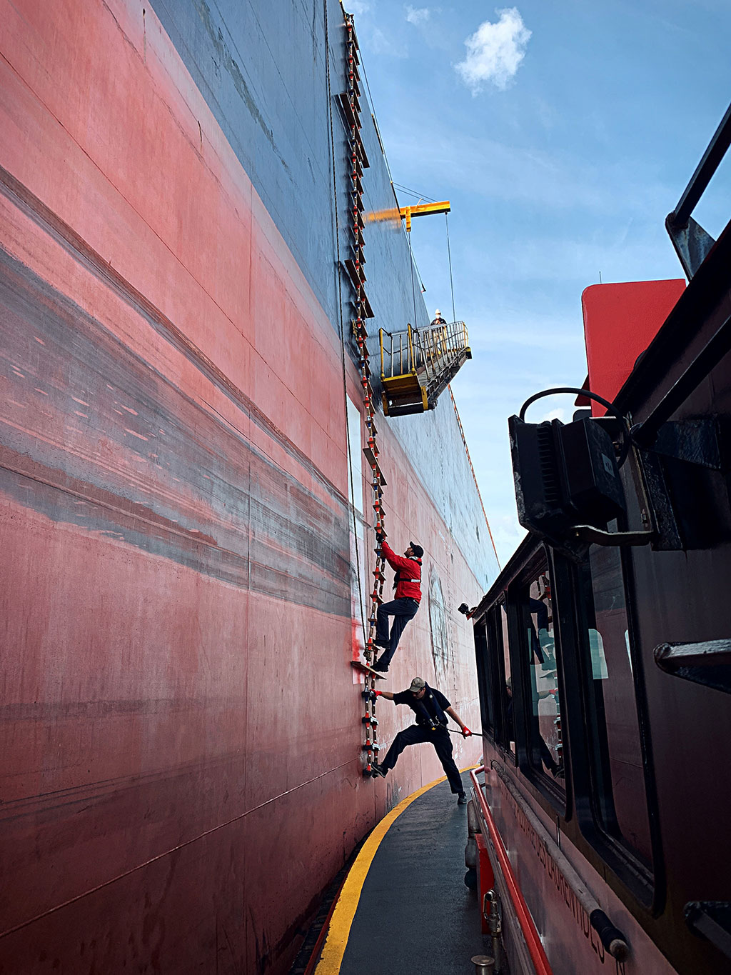 Photo of a pilot climbing up a flexible pilot ladder to an accommodation ladder resting against a ship, while a person on a pilot boat beside the ship steadies the pilot’s ladder, and a person on the ship stands by the accommodation ladder
