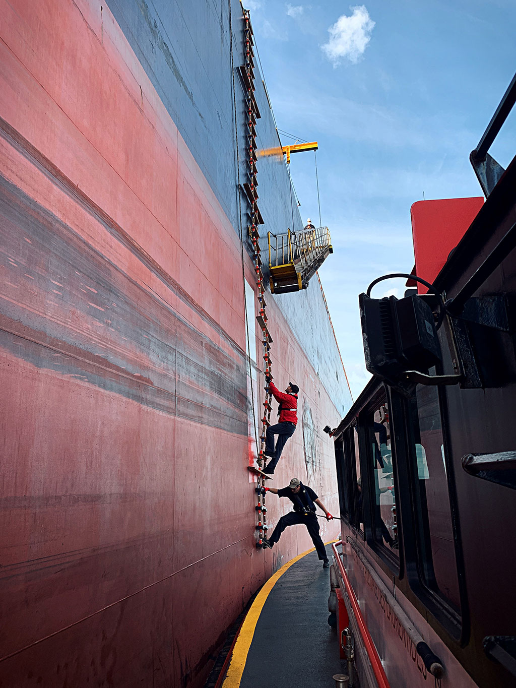 Photo montrant un pilote gravissant une échelle de pilote souple menant à une échelle de coupée fixée à un navire, tandis qu’une personne sur le bateau pilote à côté du navire stabilise l’échelle de pilote et qu’une personne sur le navire se tient près de l’échelle de coupée