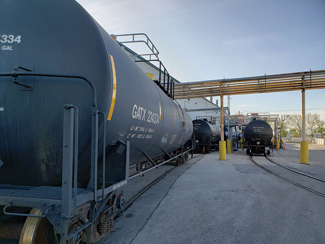Photograph of 2 sets of black tank cars sitting on 2 sets of rail tracks near a warehouse