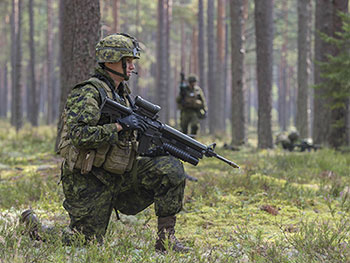 Photo of Canadian Army Reserve soldiers in a forest in Eastern Europe in 2015. In the foreground, a soldier is kneeling and holding a rifle.